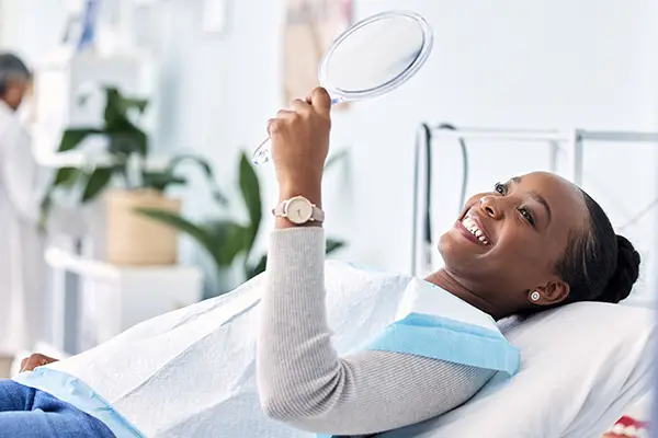Black woman holding mirror and smiling at Wasilla, AK woman happy about her new smile through cosmetic dentistry.